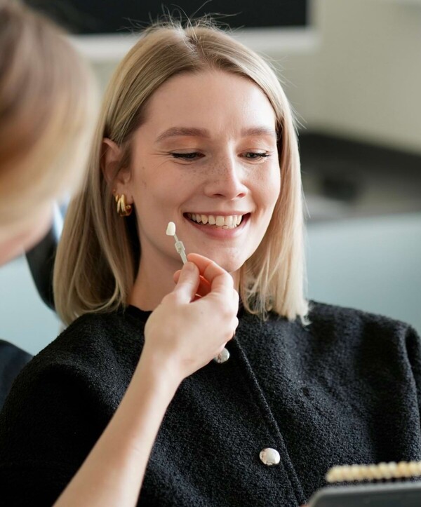 Isabell Jordan hebt einer Patientin einen Zahn aus einer Zahnfarbskala an den offen lachenden Mund.
