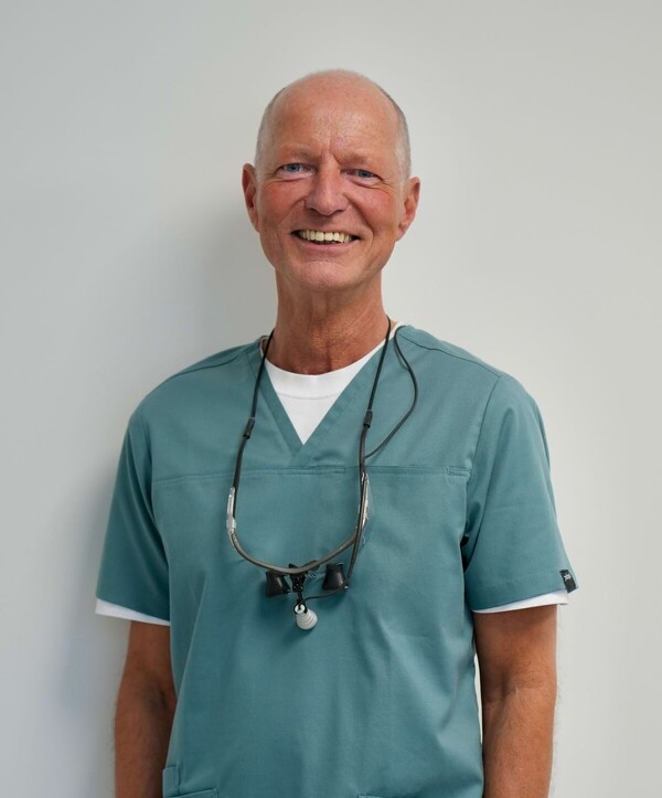 Portrait des angestellten Zahnarztes Sven Fehring bei Jordan2 in Osnabr&uuml;ck. Er steht in grau/gr&uuml;nem Kasak mit Lupenbrille um den Hals vor einer wei&szlig;en Wand.