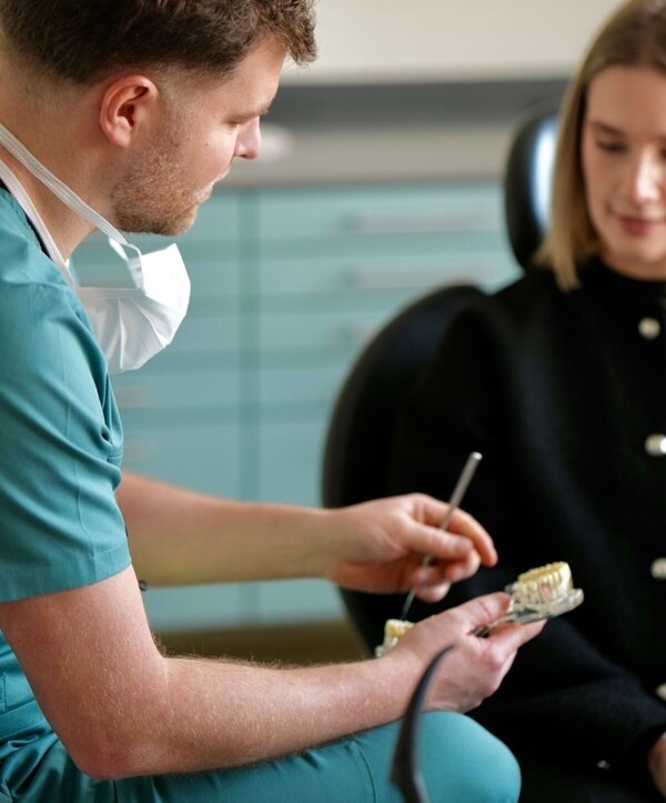 Erik Jordan h&auml;lt ein Kiefermodell in der rechten Hand und erkl&auml;rt einer Patientin mit Hilfe des Modells etwas. Die Patientin schaut konzentriert zu.