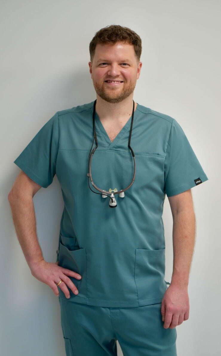 Portraitbild von Zahnarzt Erik Jordan. Er steht in gr&uuml;n/grauem Kasak vor einer wei&szlig;en Wand und st&uuml;tzt die rechte Hand auf die H&uuml;fte.