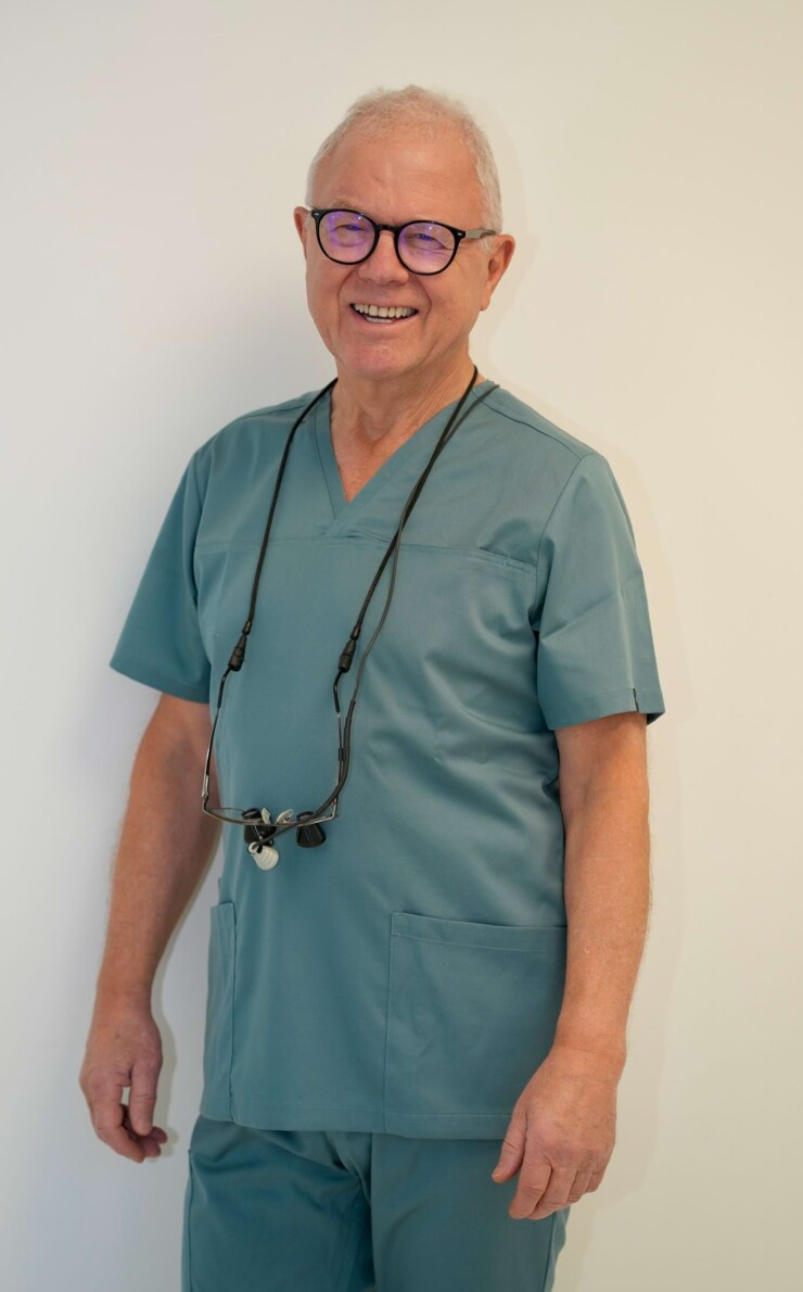 Portraitbild von Zahnarzt Wolfgang Jordan. Er steht in gr&uuml;n/grauem Kasak vor einer wei&szlig;en Wand.