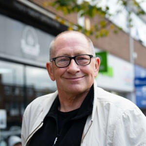 Portr&auml;t eines l&auml;chelnden, brillentragenden Mannes mit sehr kurzem Haar, der eine helle Jacke &uuml;ber einem schwarzen Oberteil tr&auml;gt.