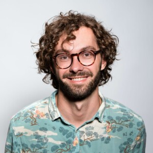 Portr&auml;t eines l&auml;chelnden Mannes mit braunem Lockenkopf, Brille und Bart, der ein hellblaues Hemd mit japanischem Muster tr&auml;gt.