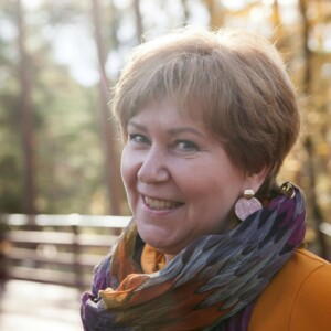 Portr&auml;t einer l&auml;chelnden Frau mit kurzen, hellbraunen Haaren, einem gemusterten Schal und gro&szlig;en Ohrringen vor einem unscharfen Waldhintergrund.