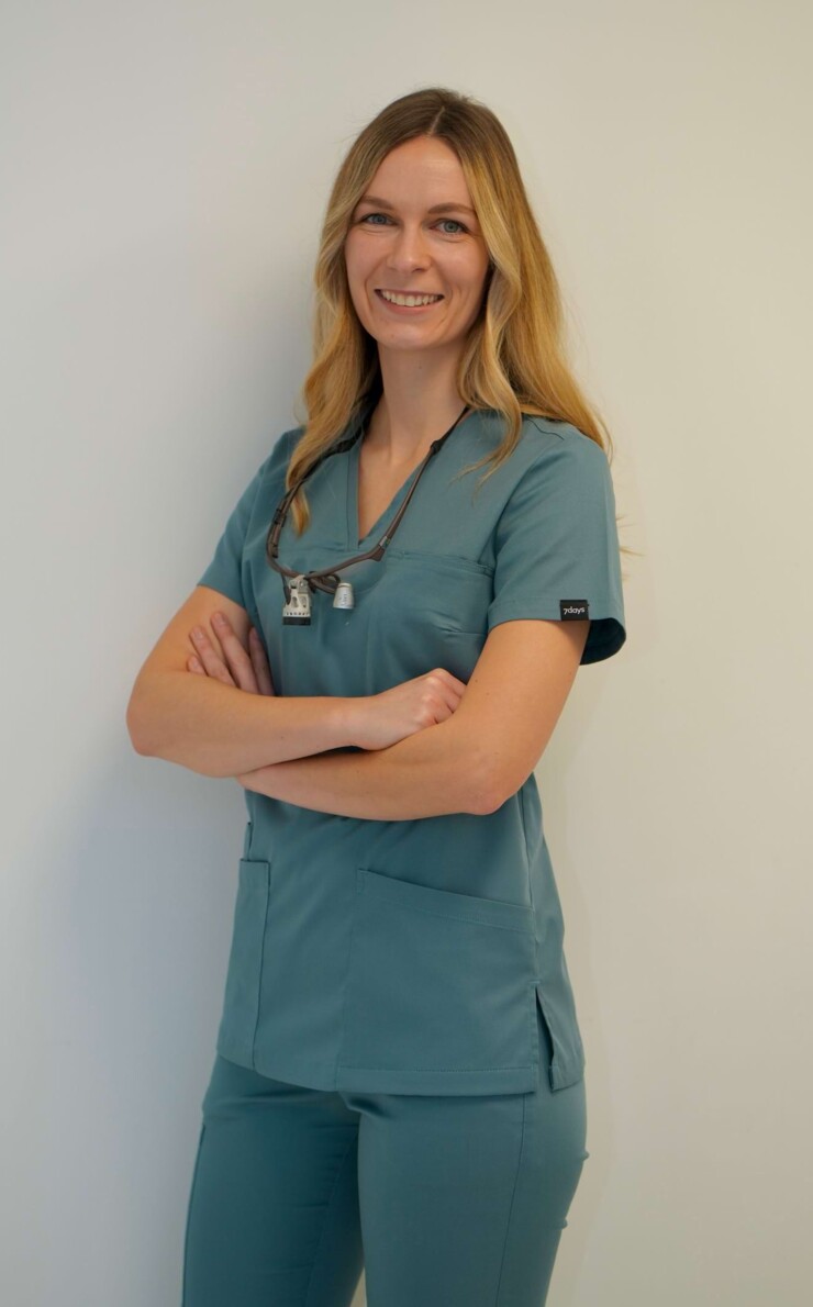 Portraitbild von Zahn&auml;rztin Isabell Jordan. Sie steht in gr&uuml;n/grauem Kasak vor einer wei&szlig;en Wand und verschr&auml;nkt die Arme.