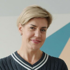 Portr&auml;t einer l&auml;chelnden Frau mit kurzen blonden Haaren, die einen dunklen Strickpullover mit wei&szlig;-blau gestreiftem V-Ausschnitt tr&auml;gt.