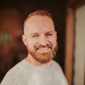 Portr&auml;t eines l&auml;chelnden Mannes mit kurzem, r&ouml;tlich-blondem Haar und Vollbart, der ein graues Oberteil tr&auml;gt.